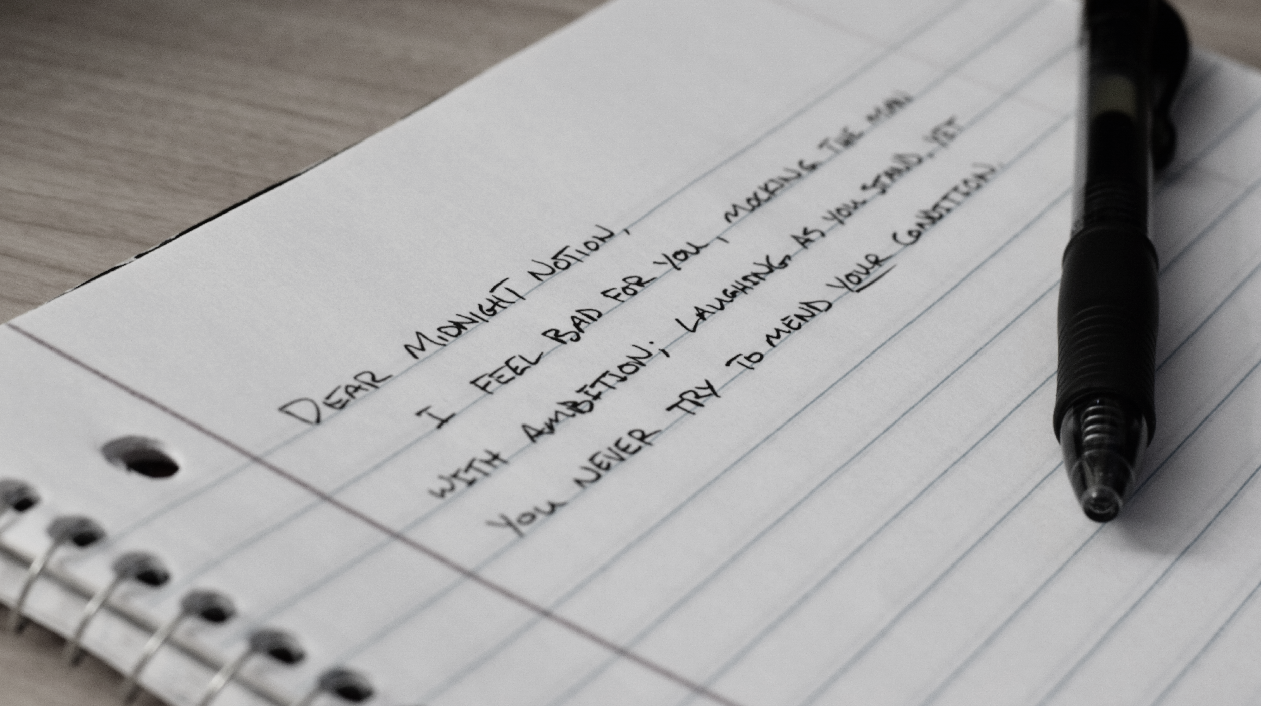 A close-up image of a lined paper notebook with a black pen lying on the page. Written on the first few lines are "Dear Midnight Notion, I feel bad for you, mocking the man with ambition; laughing as you stand, yet you never try to mend your condition."