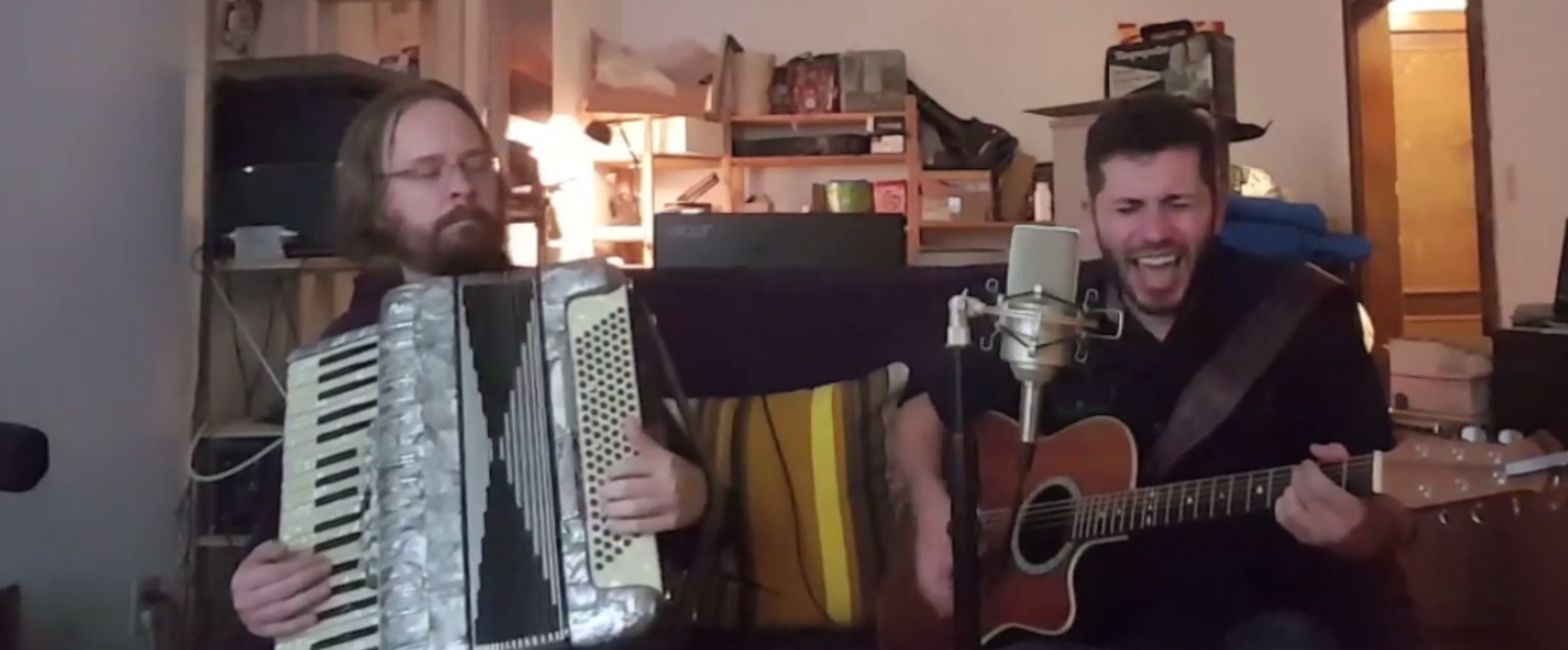 An image of Erik Ostrom and Bryce Kalal. Erik is a white man with medium-length brown hair, a beard, and glasses; Bryce is a white man with short dark brown hair and a beard. They're sitting beside each other, in front of a microphone in a messy apartment. Erik is playing a white and black accordion, while Bryce is singing and playing an acoustic guitar.