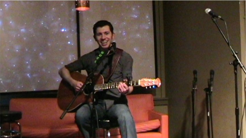 A still frame from a video of Bryce Kalal playing an acoustic guitar while sitting on a stool and singing into a microphone at Corner Coffee.