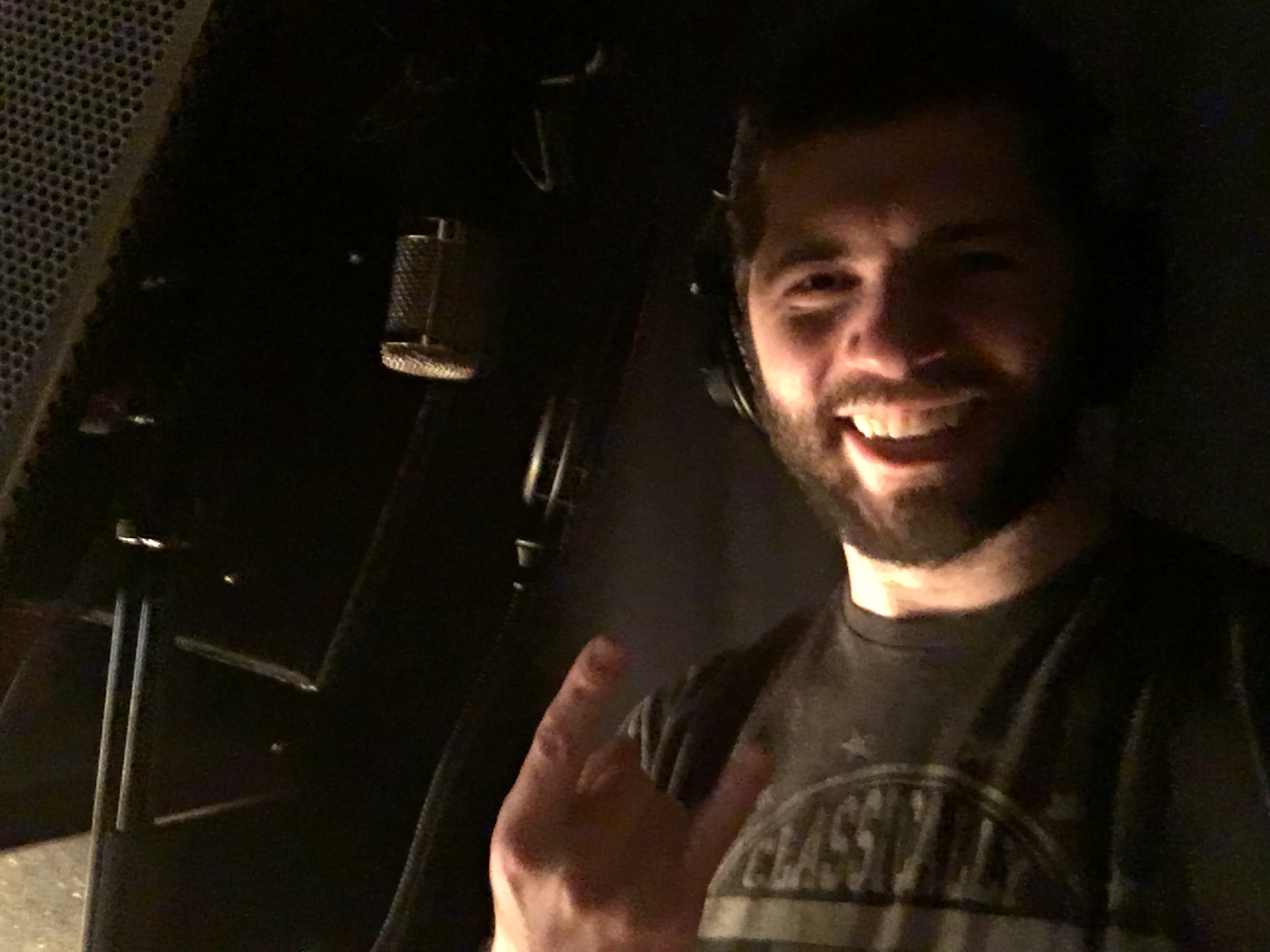 A selfie of Bryce Kalal smiling, showing "the horns" with his hand, and standing next to a microphone in a vocal recording booth.
