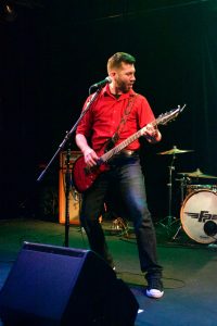 Bryce Kalal playing a red guitar on stage at The Garage. He is wearing a red button-up shirt with the sleeves rolled up, and black jeans.