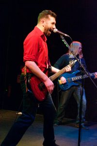 Bryce Kalal singing into a microphone and playing a red guitar on stage at The Garage. He is wearing a red button-up shirt with the sleeves rolled up, and black jeans. In the background, Evan Paetzel is playing a blue bass guitar.