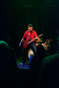 Bryce Kalal singing into a microphone and playing a red guitar on stage at The Garage. He is wearing a red button-up shirt with the sleeves rolled up, and black jeans. In the foreground is the back side of several audience people, and in the background is David Koepplin Jr. playing the drums.