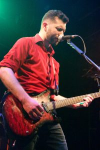 Bryce Kalal singing into a microphone and playing a red guitar on stage at The Garage. He is wearing a red button-up shirt with the sleeves rolled up, black jeans, and his eyes are tightly closed as he sings.