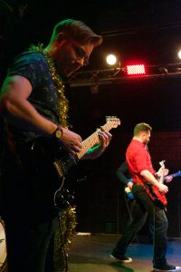 Chris Johnson is playing a black guitar on stage at The Garage. He's wearing glasses, a tight-fitting blue shirt, and has a golden garland draped around his neck. In the background, Bryce Kalal is playing a red guitar while wearing a red button-up shirt with the sleeves rolled up and black jeans.