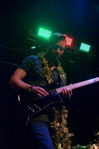 Chris Johnson is playing a black guitar on stage. He's wearing glasses, a tight-fitting blue shirt, and has a golden garland draped around his neck.