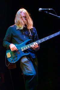 Evan Paetzel is playing a bass guitar on stage. He's wearing glasses, a long-sleeved blue button-up shirt, and jeans. His long blonde hair is in his face as he plays.