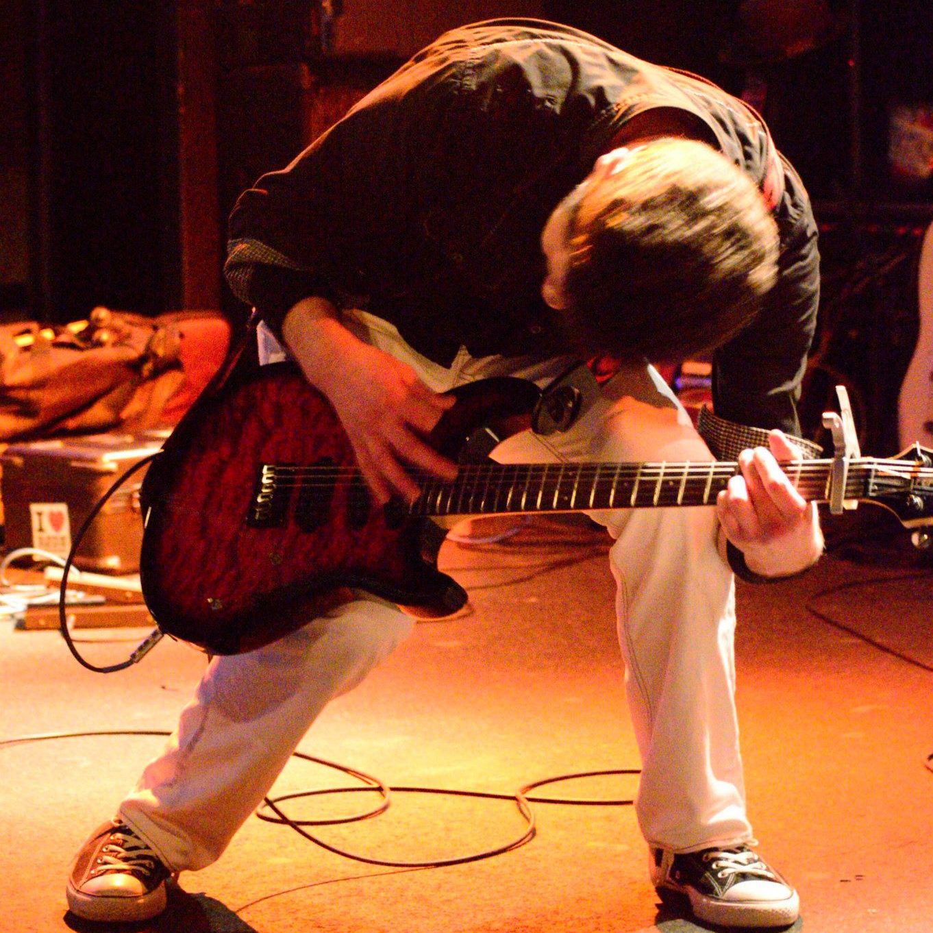 An image of Bryce Kalal, a white man with a black shirt and white pants, hunched over as he plays a red guitar on stage.